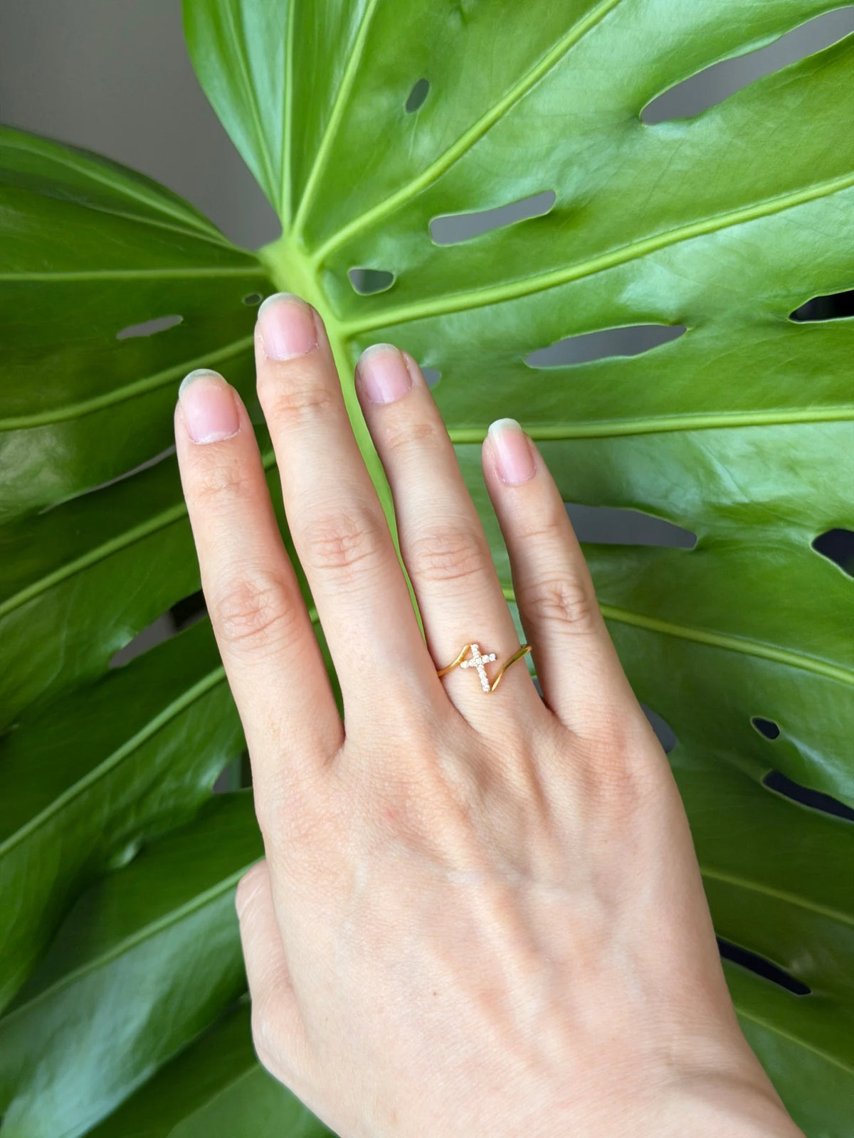 Hand wearing the dainty gold cross ring against a green monstera leaf